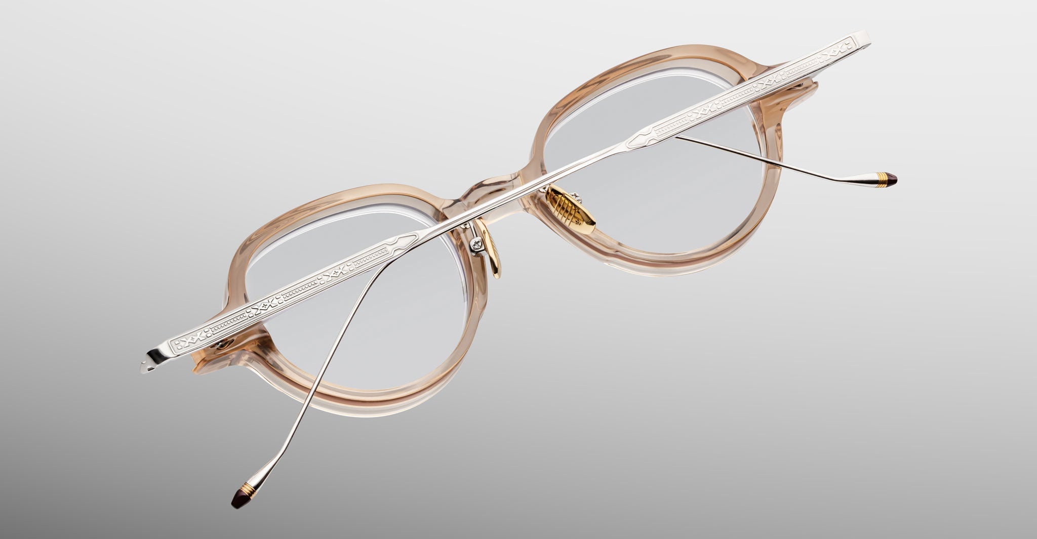The Jacques Marie Mage Clark eyeglasses feature clear frames, metal arms, and decorative temple detailing. Handcrafted in Japan, they are displayed on a light gradient background.