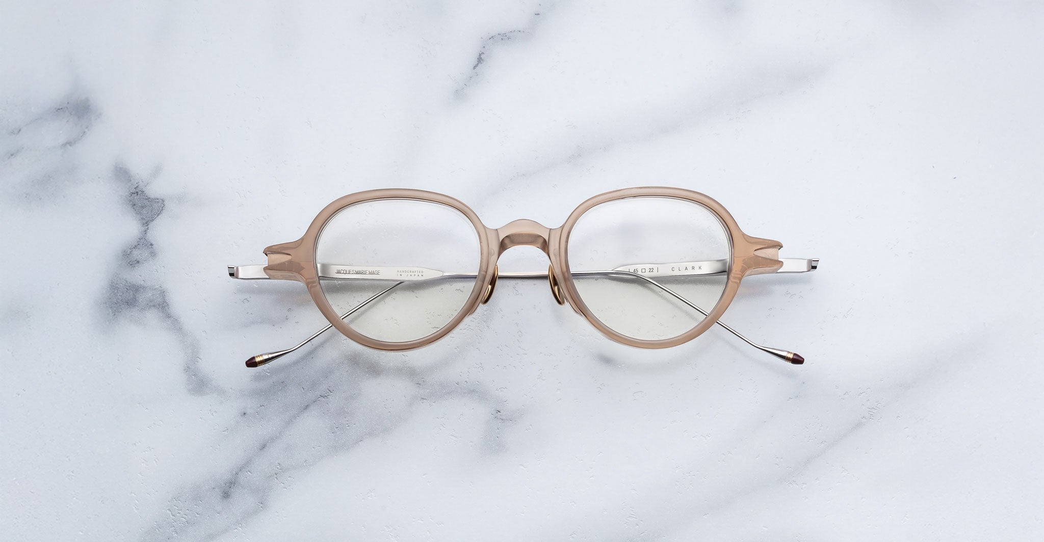 Jacques Marie Mage Clark eyeglasses by Jacques Marie Mage, handcrafted in Japan with a beige round frame and clear lenses, displayed on a white marble surface.