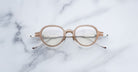 Jacques Marie Mage Clark eyeglasses by Jacques Marie Mage, handcrafted in Japan with a beige round frame and clear lenses, displayed on a white marble surface.