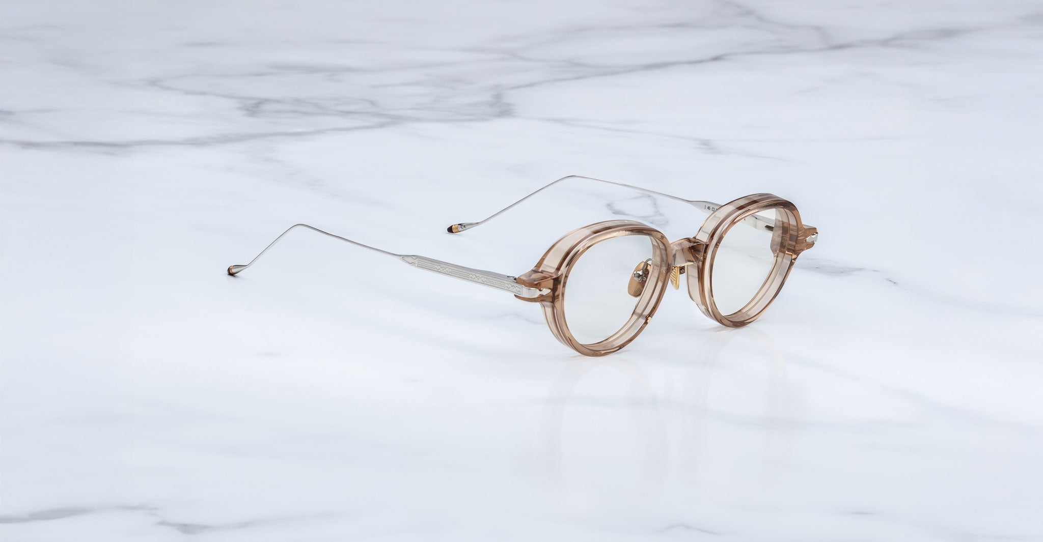 Round, translucent eyeglasses with thin metal arms rest on a white marble surface—midcentury-inspired Jacques Marie Mage Clark optical frames.