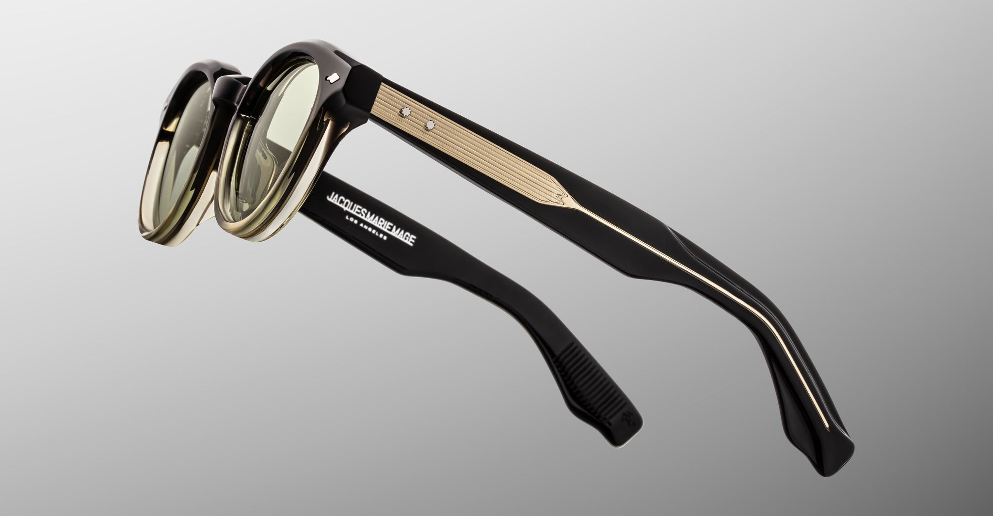 The Jacques Marie Mage Clemens eyewear in black and gold with green-tinted lenses is displayed from a rear side angle on a gray background. "Jacques Marie Mage" is printed in white on the inside of the left temple arm.