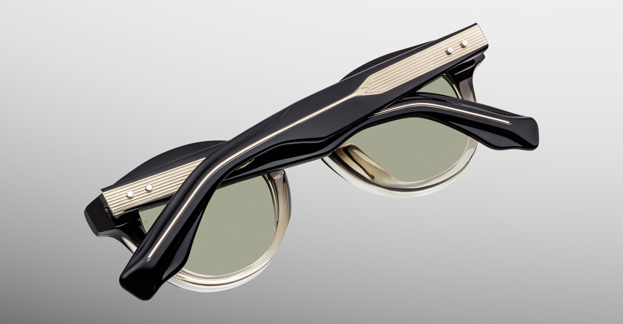 A pair of Jacques Marie Mage Clemens black and gold sunglasses, folded closed and viewed from above on a light gray background.