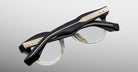 A pair of Jacques Marie Mage Clemens eyeglasses in black with thick, sculpted frames and gold accents on the temples, displayed against a light gray background.