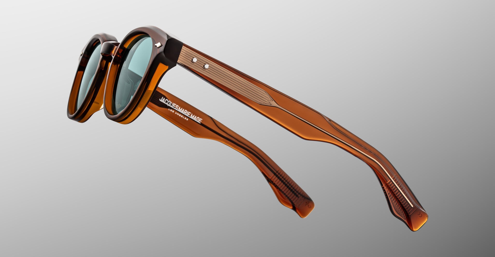 The Jacques Marie Mage Clemens sunglasses feature brown acetate Wellington frames, green tinted lenses, silver rivets, and the brand’s logo on the temple. This limited-edition eyewear is set against a neutral gradient background.