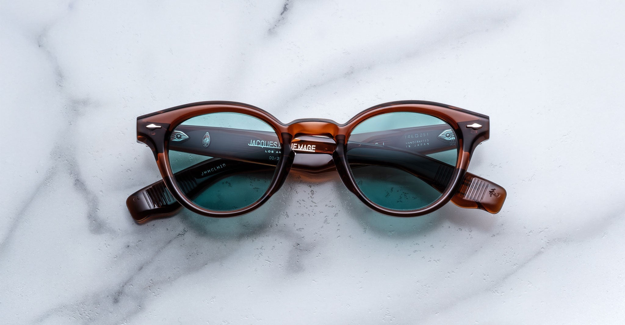 Jacques Marie Mage Clemens sunglasses featuring round brown frames and blue-tinted lenses, displayed on a white marble surface.