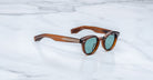 Jacques Marie Mage Clemens sunglasses in brown tortoiseshell with green lenses and thick arms rest on a white marble surface. These limited-edition frames feature silver decorative details on the arms.