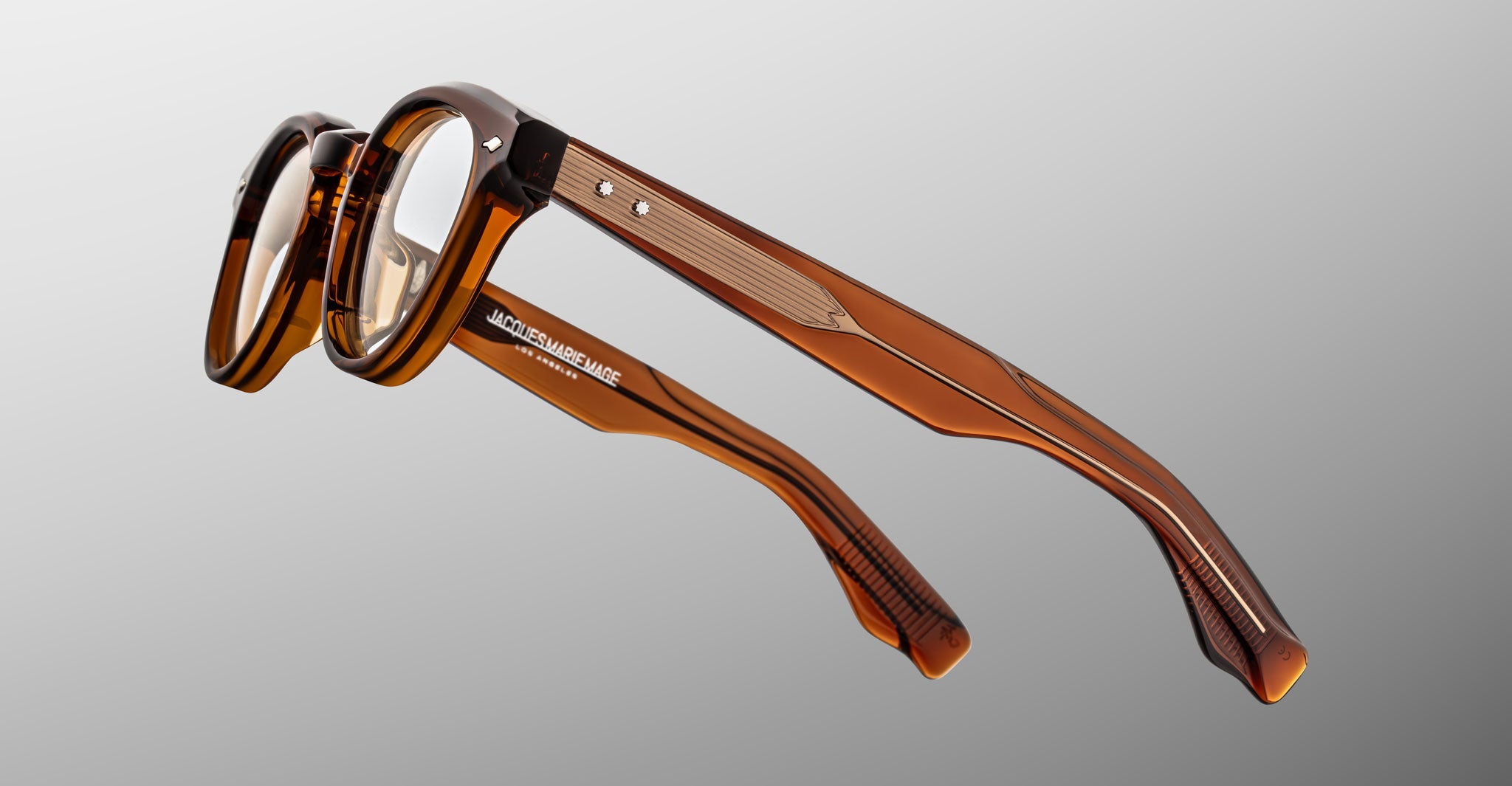 The Jacques Marie Mage Clemens eyeglasses by Jacques Marie Mage feature brown transparent thick frames and visible silver hinge details, shown against a plain grey background.