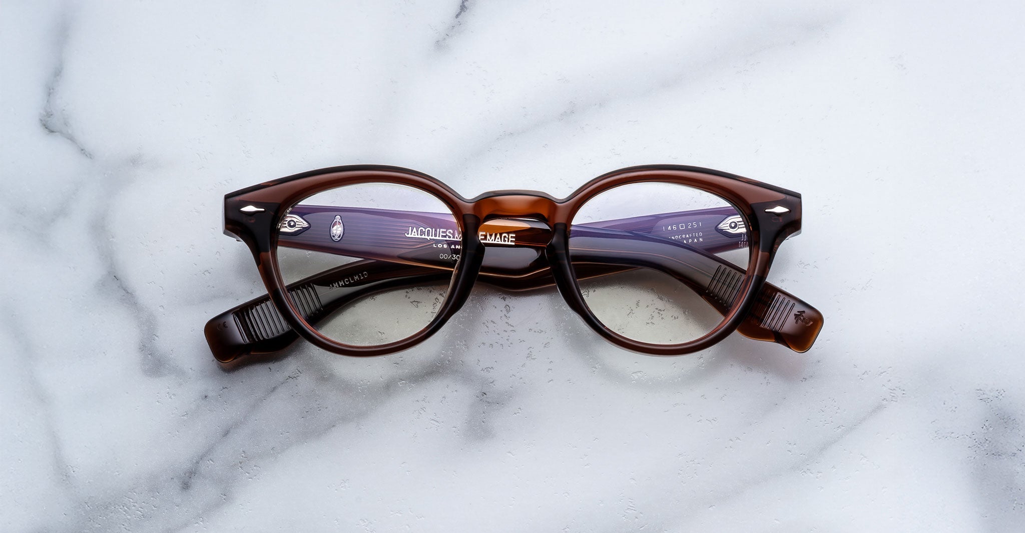 A pair of Jacques Marie Mage Clemens round brown eyeglasses with clear lenses rests on a white marble surface.