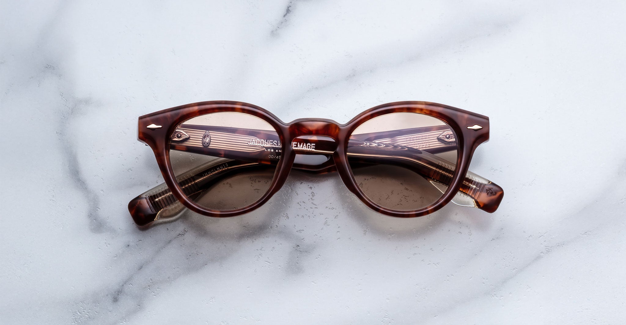 Jacques Marie Mage Clemens brown round eyeglasses with clear lenses and a subtle cat-eye shape, inspired by Wellington eyewear, are placed on a white marble surface with the arms folded inward.