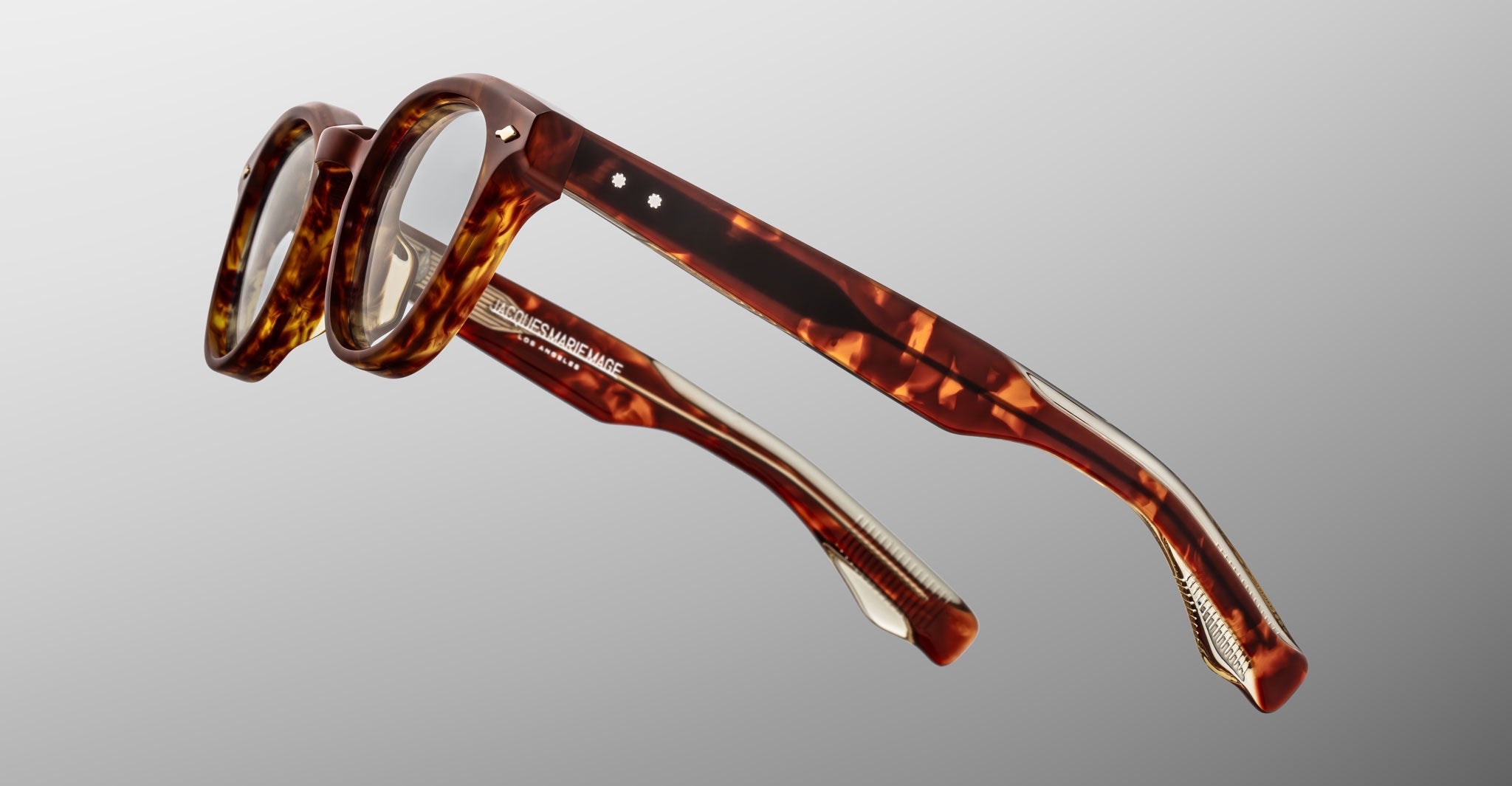 Jacques Marie Mage Clemens eyeglasses by Jacques Marie Mage feature thick round brown tortoiseshell frames and metal temple details, shown on a gray gradient background—an elevated spin on classic Wellington style.