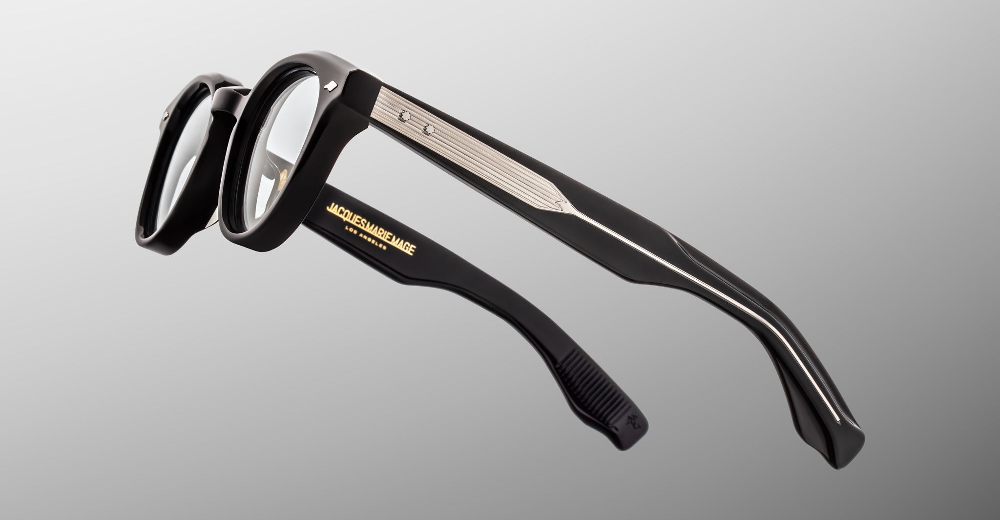 The Jacques Marie Mage Clemens eyeglasses feature black frames, clear lenses, metallic temple accents, and gold branding on the inner arm, offering a refined Wellington style displayed against a gray background.