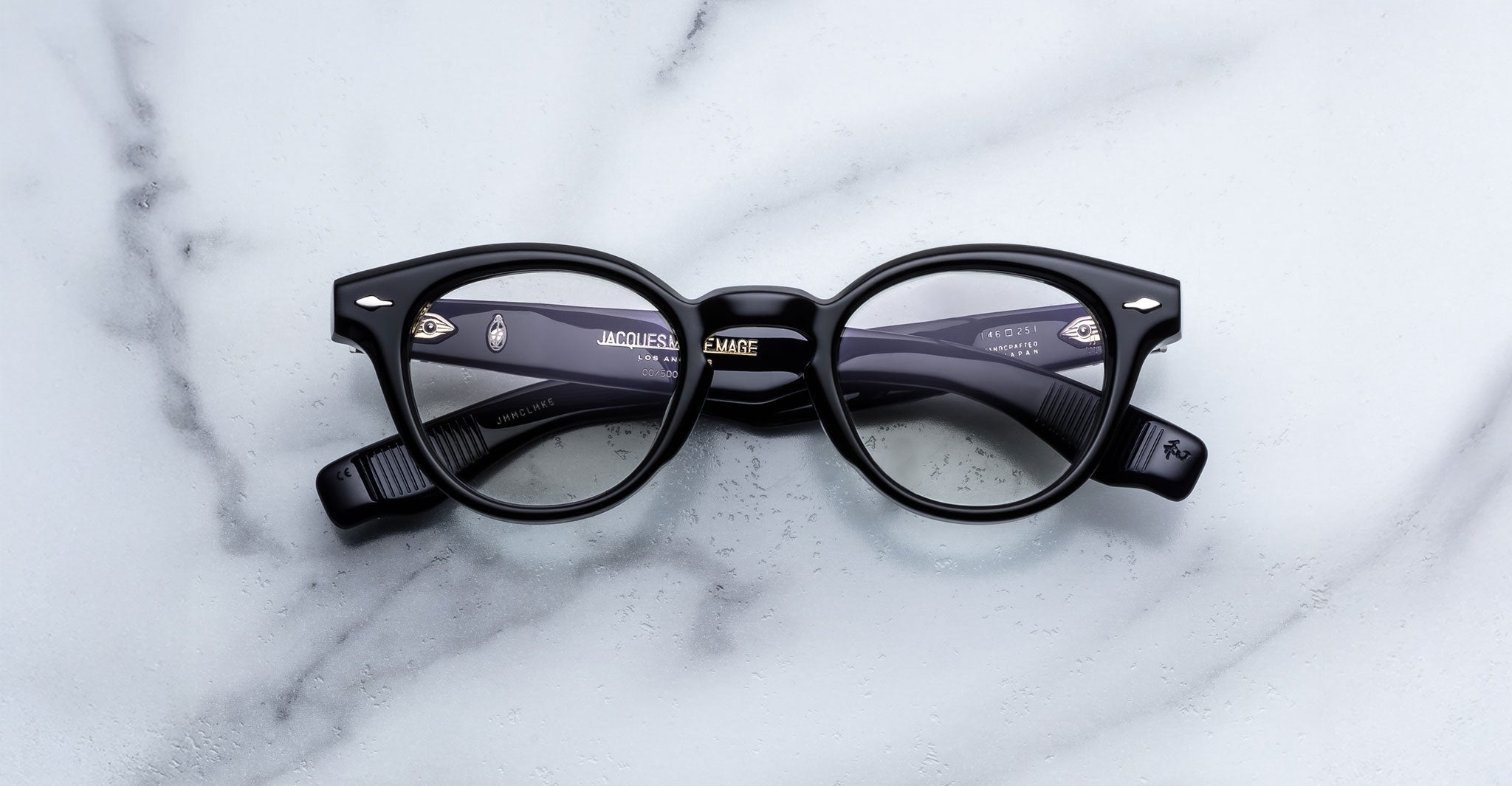 The Jacques Marie Mage Clemens by Jacques Marie Mage, featuring black round thick frames inspired by Wellington eyewear, is displayed on a white marble surface.