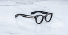 The Jacques Marie Mage Clemens features black round eyeglasses with thick frames and metal temple detailing, inspired by Wellington eyewear, displayed stylishly on a white marble surface.
