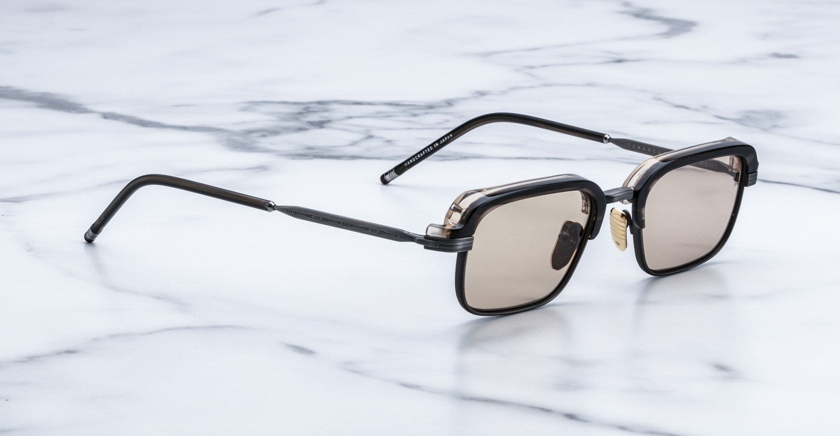 A pair of Jacques Marie Mage Comanche black square-framed sunglasses with brown tinted lenses rests on a white marble surface.