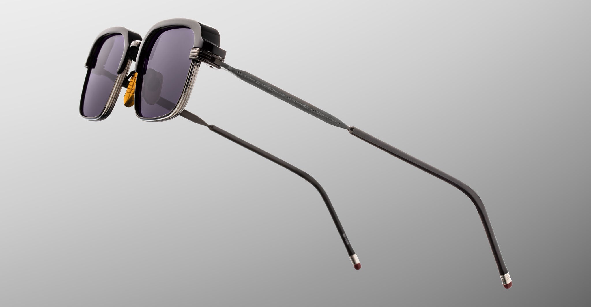 Jacques Marie Mage Comanche sunglasses—black rectangular frames with metallic accents and dark lenses—float against a neutral gray background, highlighting this limited-edition piece from Jacques Marie Mage.