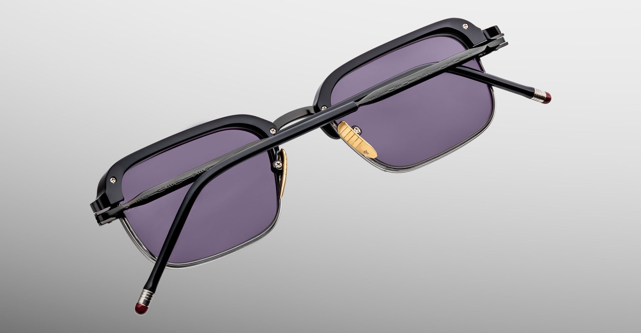 The Jacques Marie Mage Comanche sunglasses, featuring black frames, purple lenses, and gold nose pads, are displayed from the back on a light gray background.