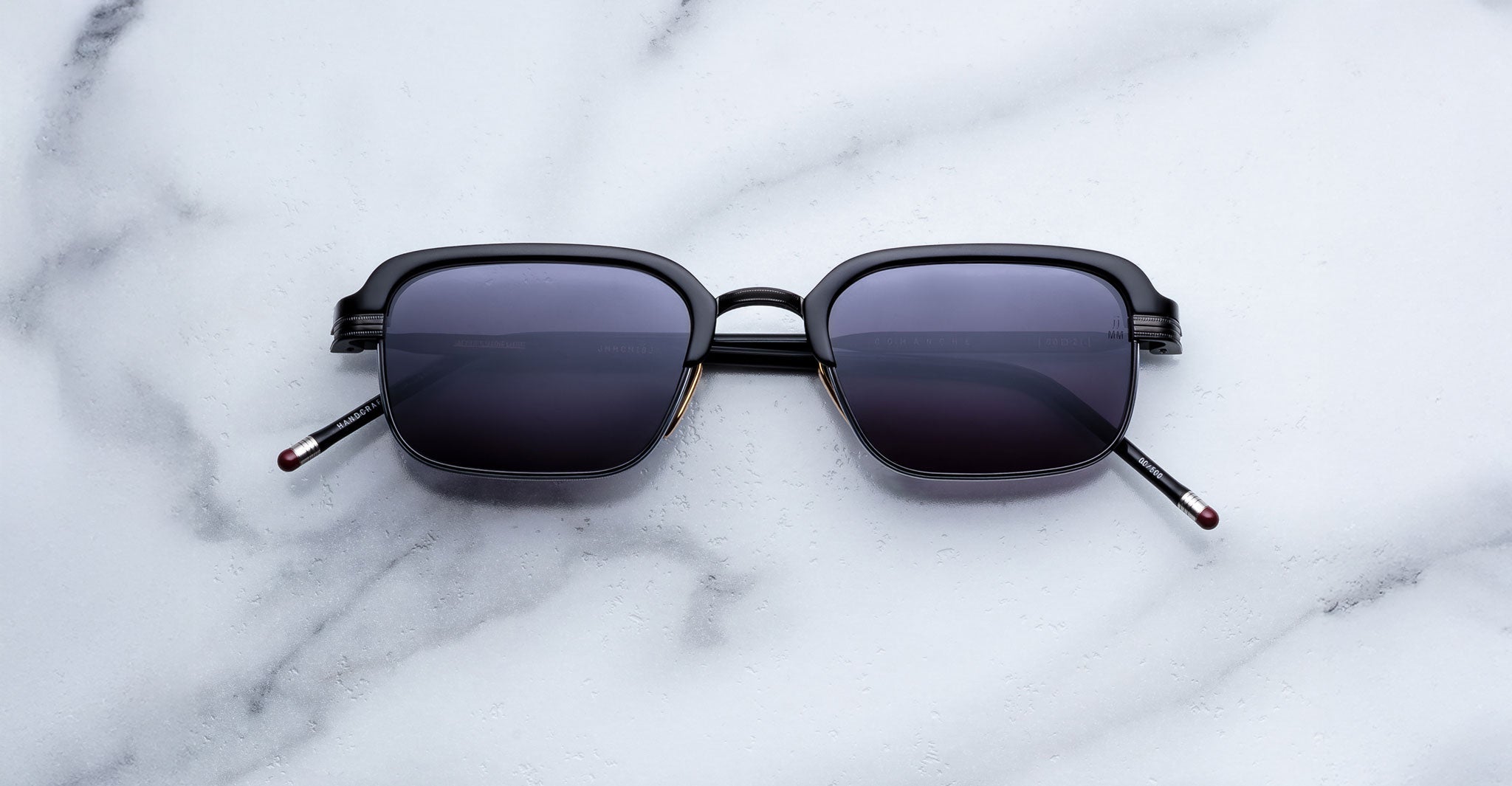 The Jacques Marie Mage Comanche sunglasses, featuring a rectangular black frame and dark lenses, rest on white marble—highlighting this limited-edition handcrafted eyewear by Jacques Marie Mage.