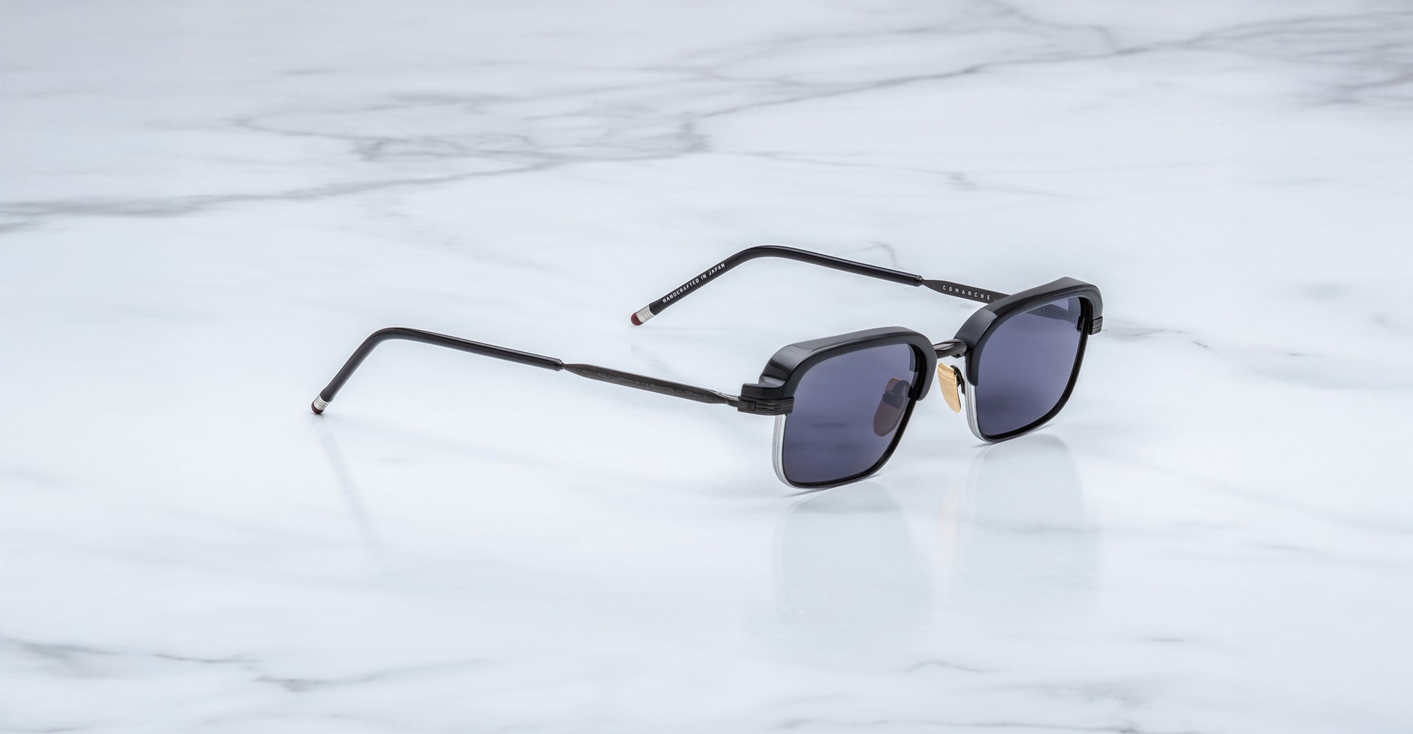 The Jacques Marie Mage Comanche, black square-frame handcrafted sunglasses with dark lenses, rests on a white marble surface.