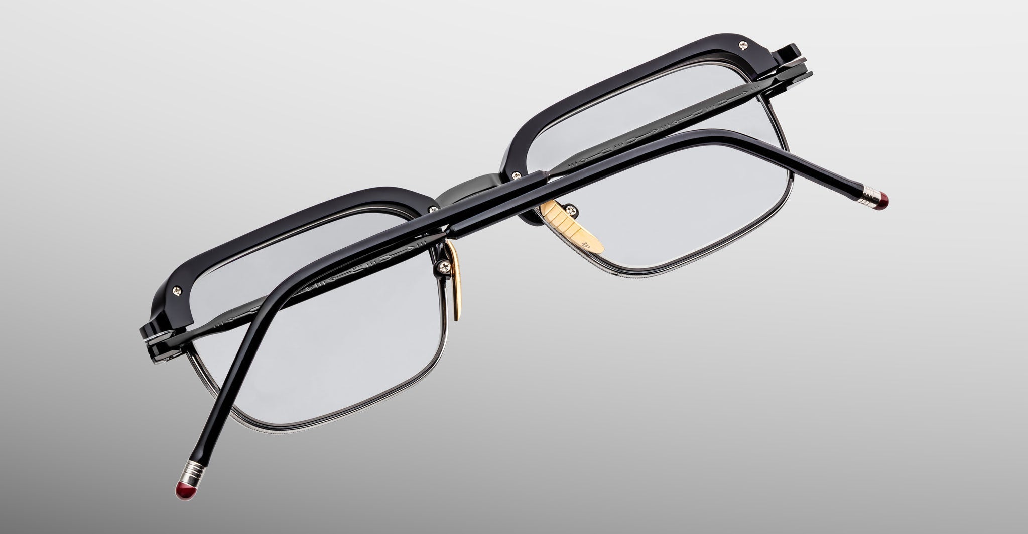 The Jacques Marie Mage Comanche is a limited-edition rectangular black eyeglass frame with clear lenses, gold nose pads, and red-tipped black arms, shown from the back against a gradient gray background.