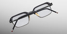 The Jacques Marie Mage Comanche is a limited-edition rectangular black eyeglass frame with clear lenses, gold nose pads, and red-tipped black arms, shown from the back against a gradient gray background.