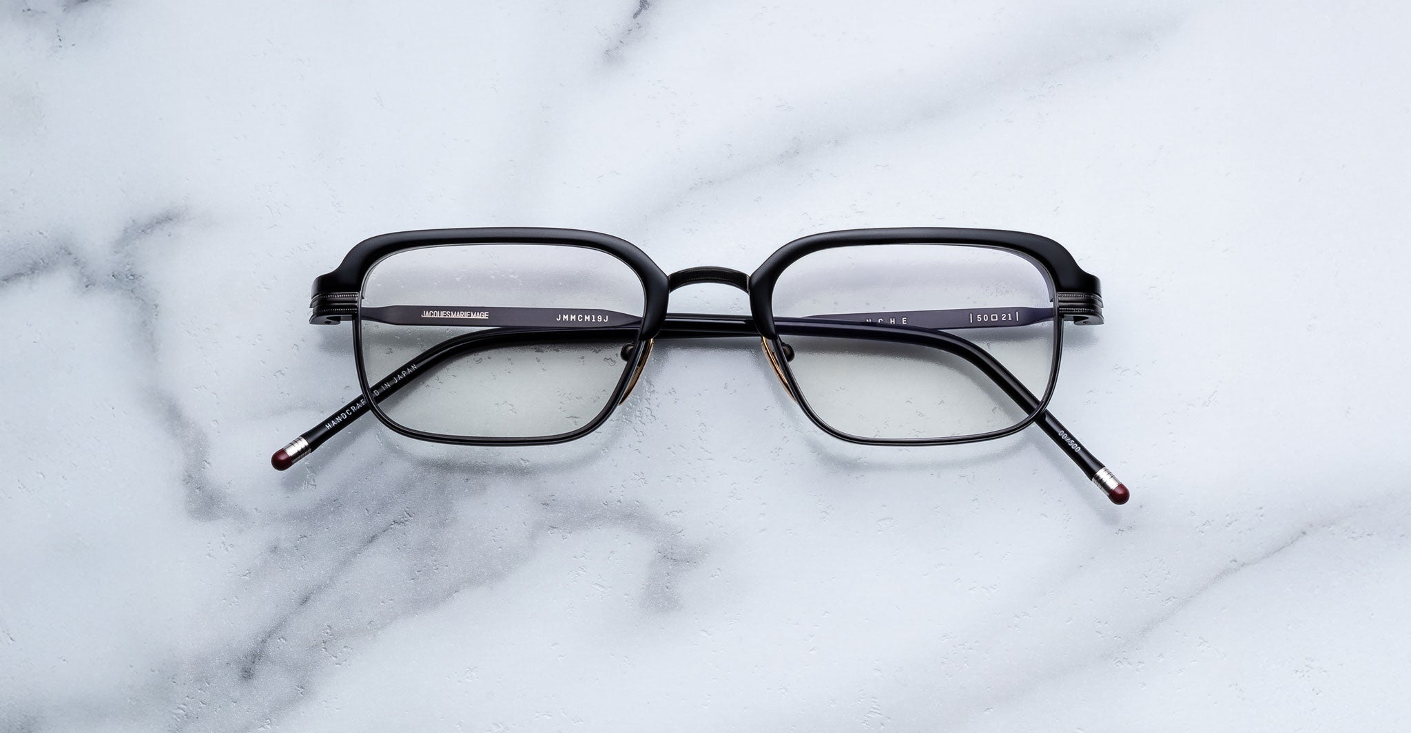 A pair of black rectangular Jacques Marie Mage Comanche eyeglasses with thin arms rests on a white marble surface, reflecting the refined elegance of this limited-edition Jacques Marie Mage eyewear.
