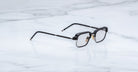 A pair of Jacques Marie Mage Comanche black rectangular eyeglasses with metal temples rests on a white marble surface, showcasing the brand’s limited-edition craftsmanship.