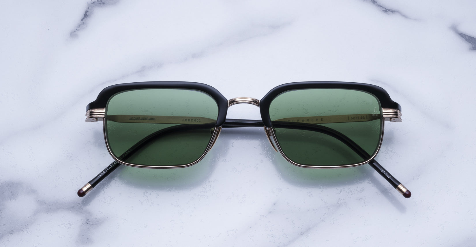 The Jacques Marie Mage Comanche sunglasses, featuring square black frames and green lenses, are displayed on a white marble surface.