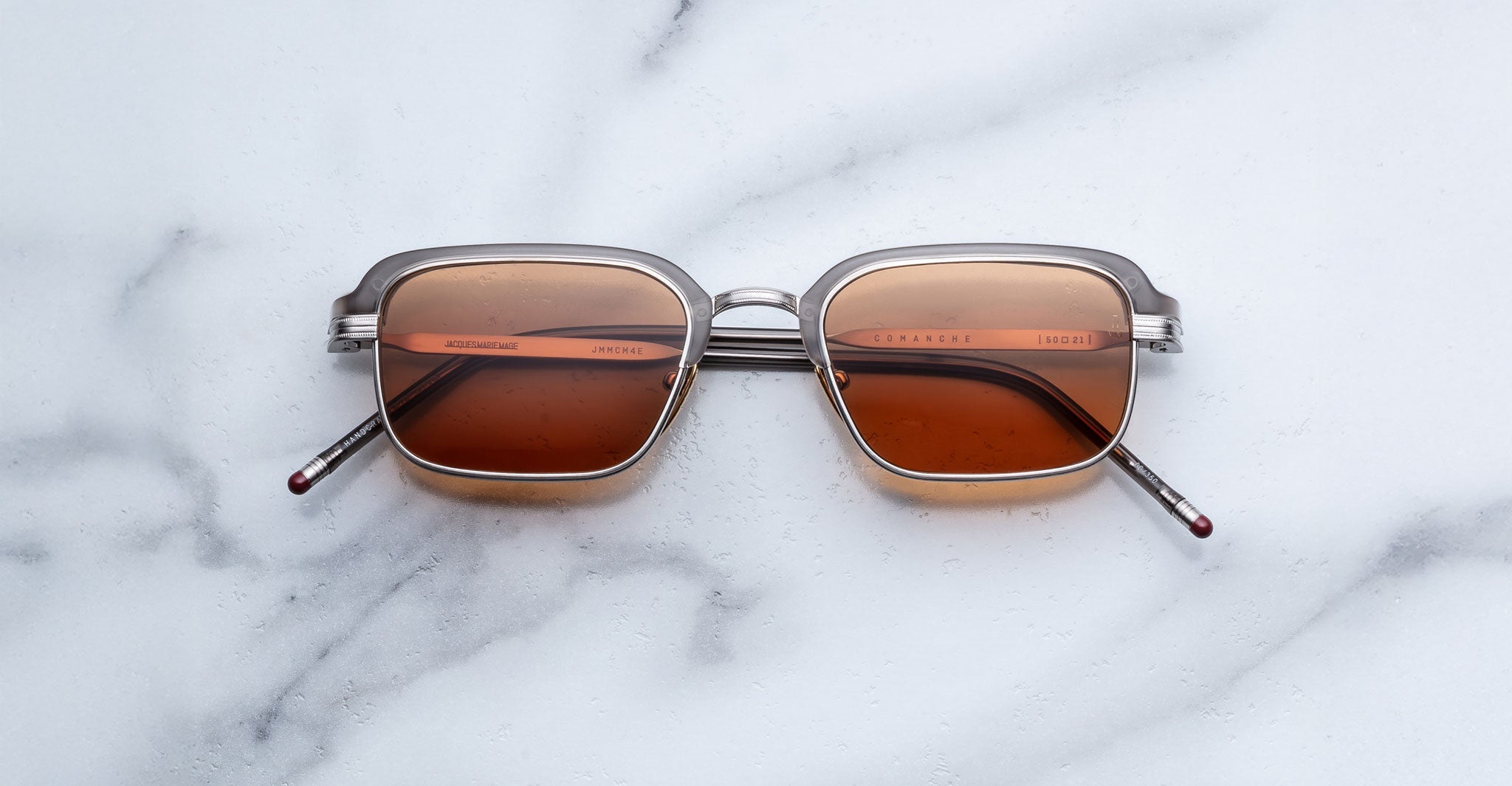 The Jacques Marie Mage Comanche sunglasses by Jacques Marie Mage feature rectangular silver frames and brown tinted lenses, showcased on a white marble surface.
