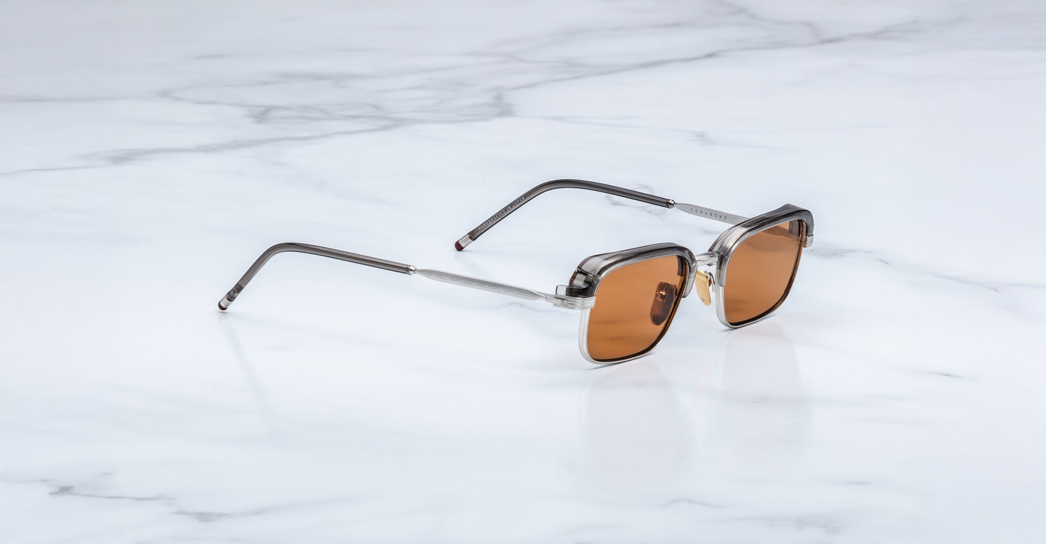 The Jacques Marie Mage Comanche sunglasses, featuring brown lenses and transparent gray frames, rest on a white marble surface—a striking limited-edition piece from Jacques Marie Mage.