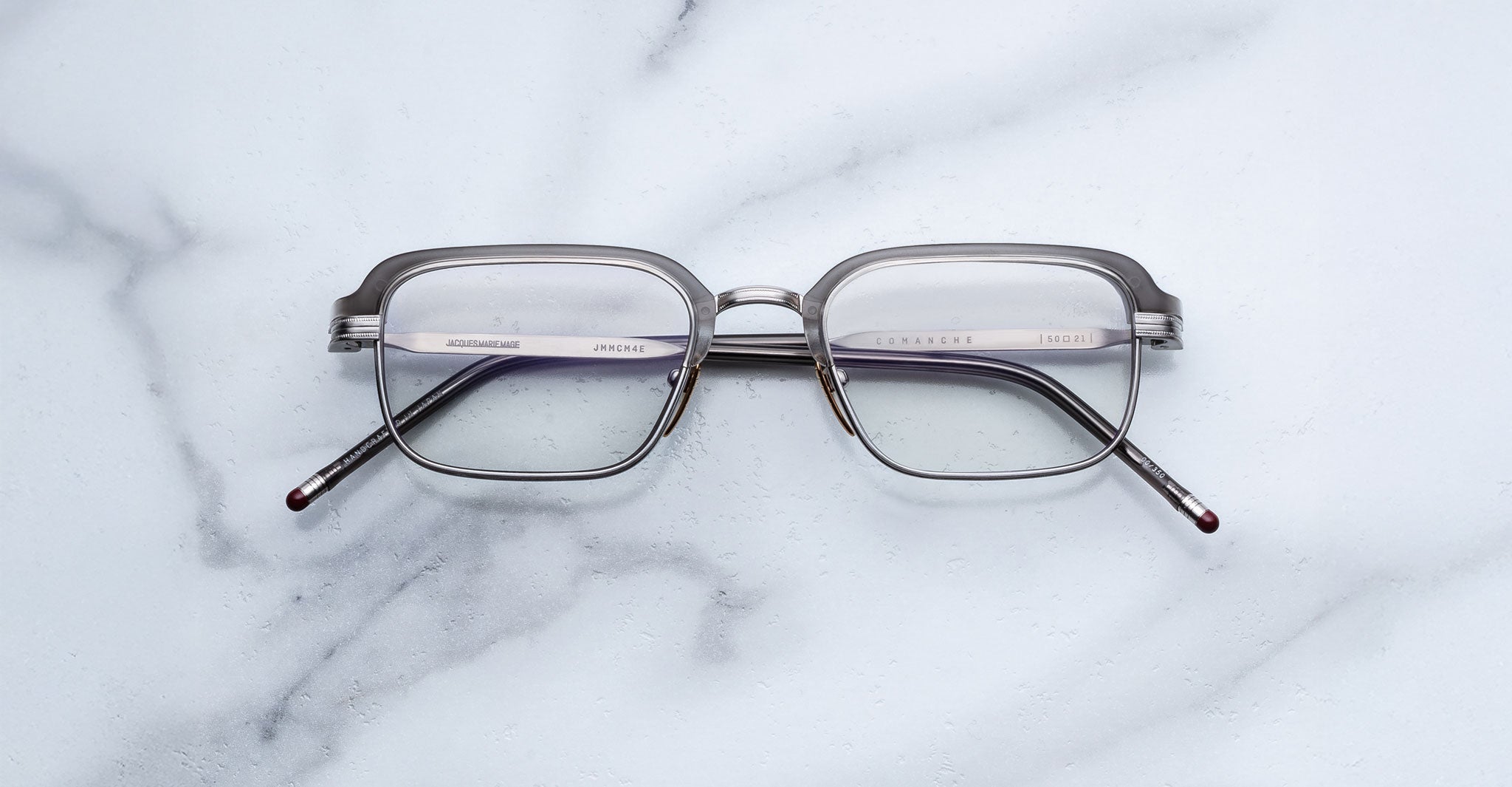 The Jacques Marie Mage Comanche eyeglasses, featuring rectangular metal frames, rest on a white marble surface, highlighting the brand's iconic limited-edition style.