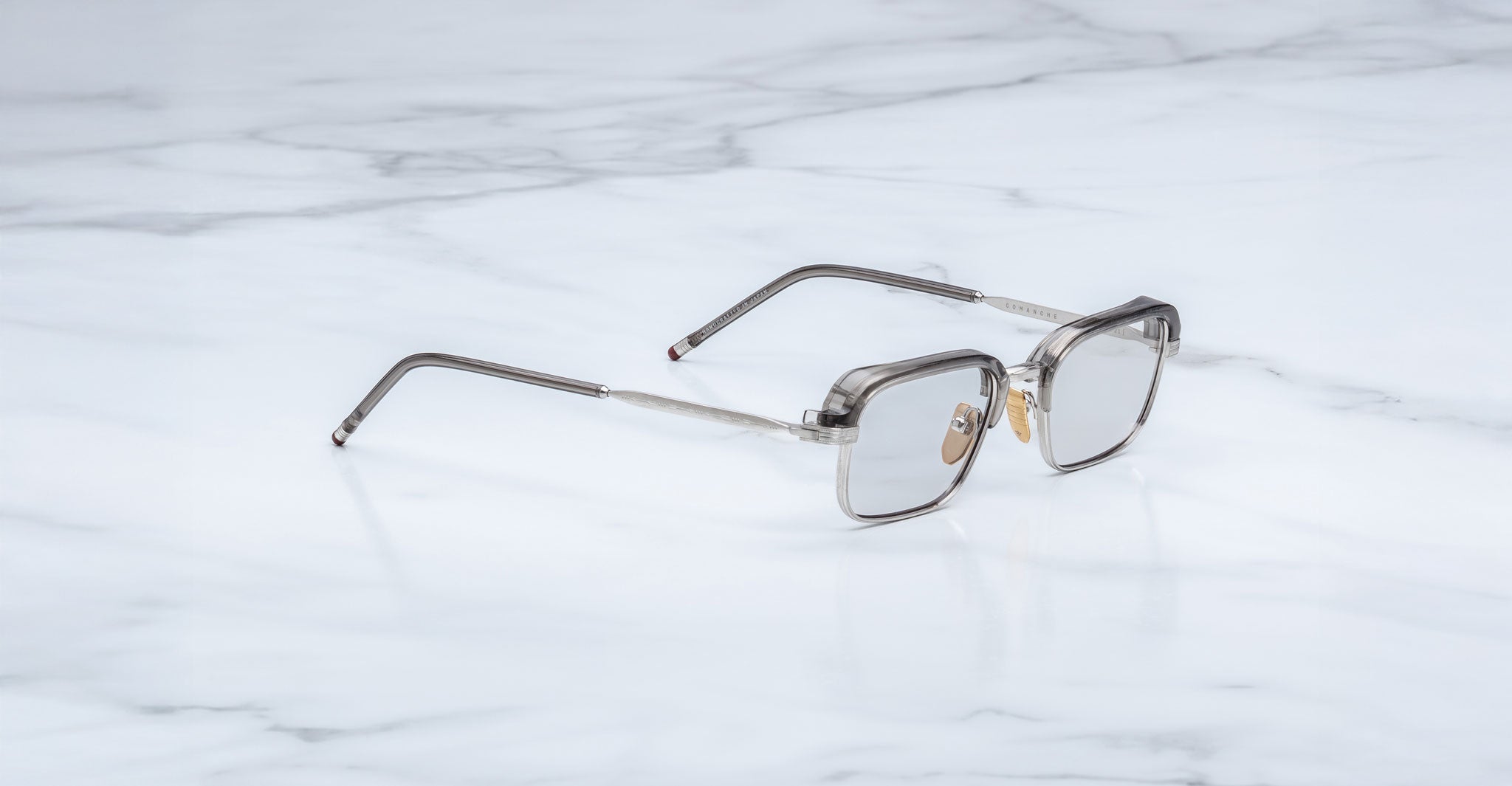 A pair of rectangular, translucent Jacques Marie Mage Comanche eyeglasses with thin arms rests on a white marble surface—a limited-edition design from Jacques Marie Mage.