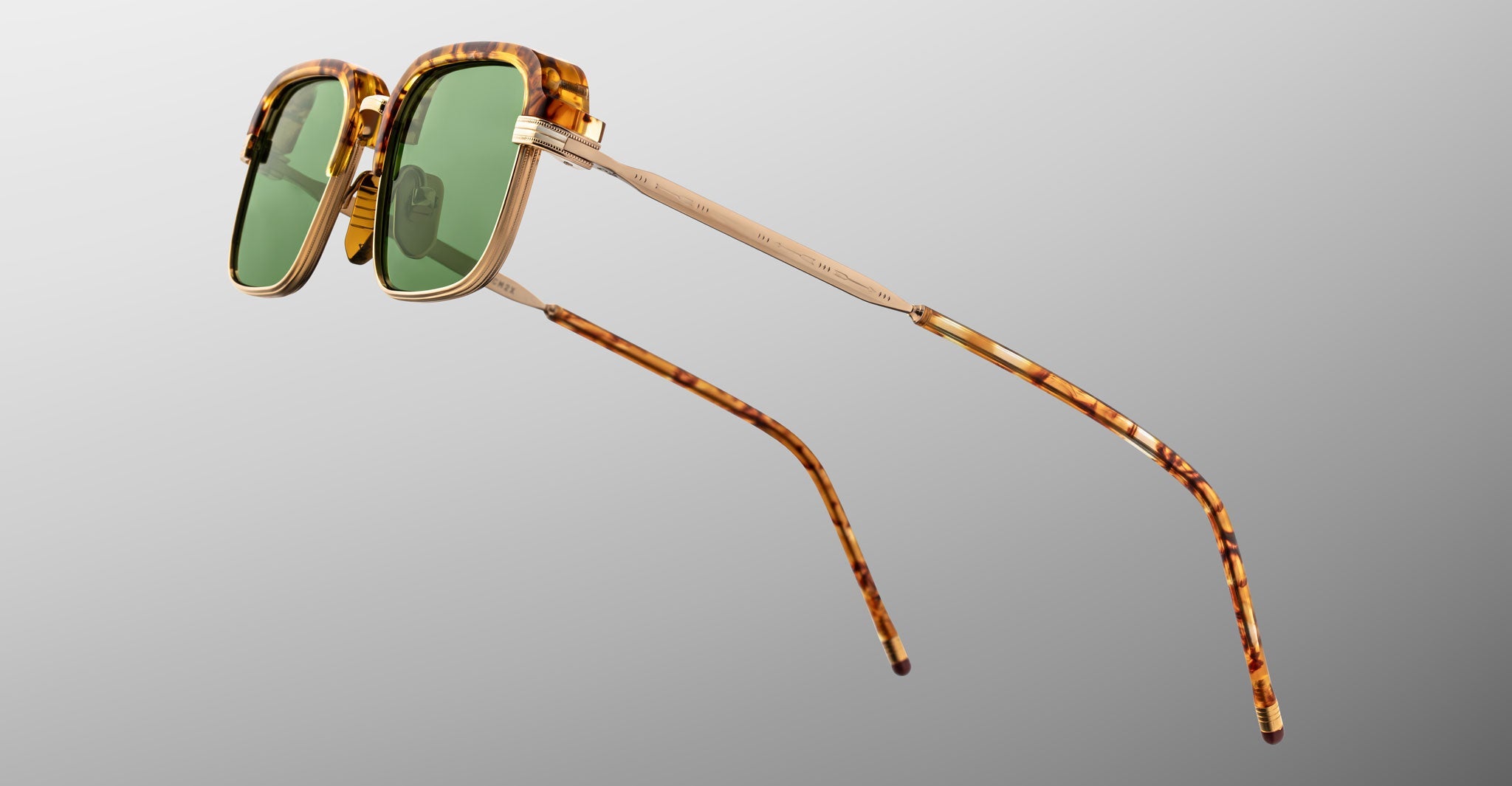 The Jacques Marie Mage Comanche sunglasses feature handcrafted tortoiseshell frames, green lenses, and gold metal accents, showcasing limited-edition style from Jacques Marie Mage against a plain grey background.