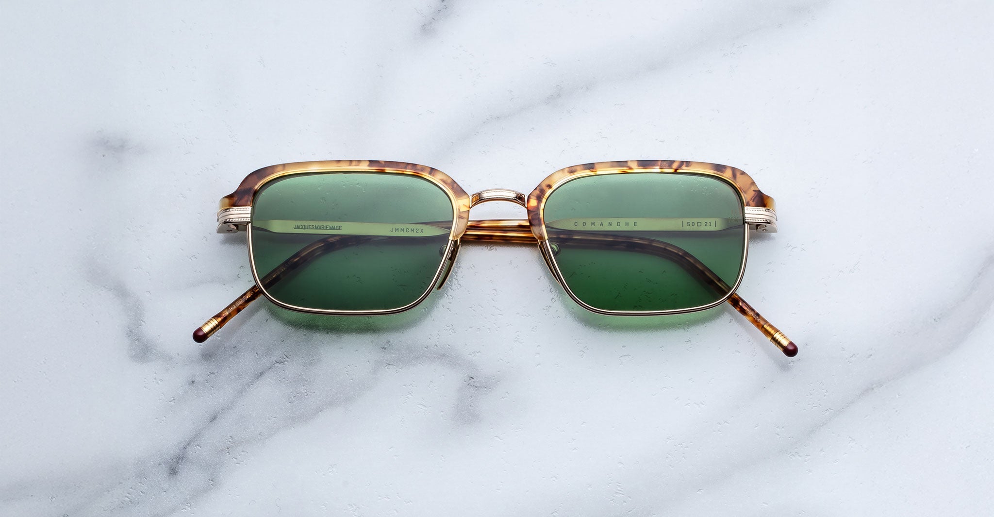 Jacques Marie Mage Comanche sunglasses by Jacques Marie Mage: limited-edition eyewear with tortoiseshell frames and green lenses, displayed flat on a white marble surface.