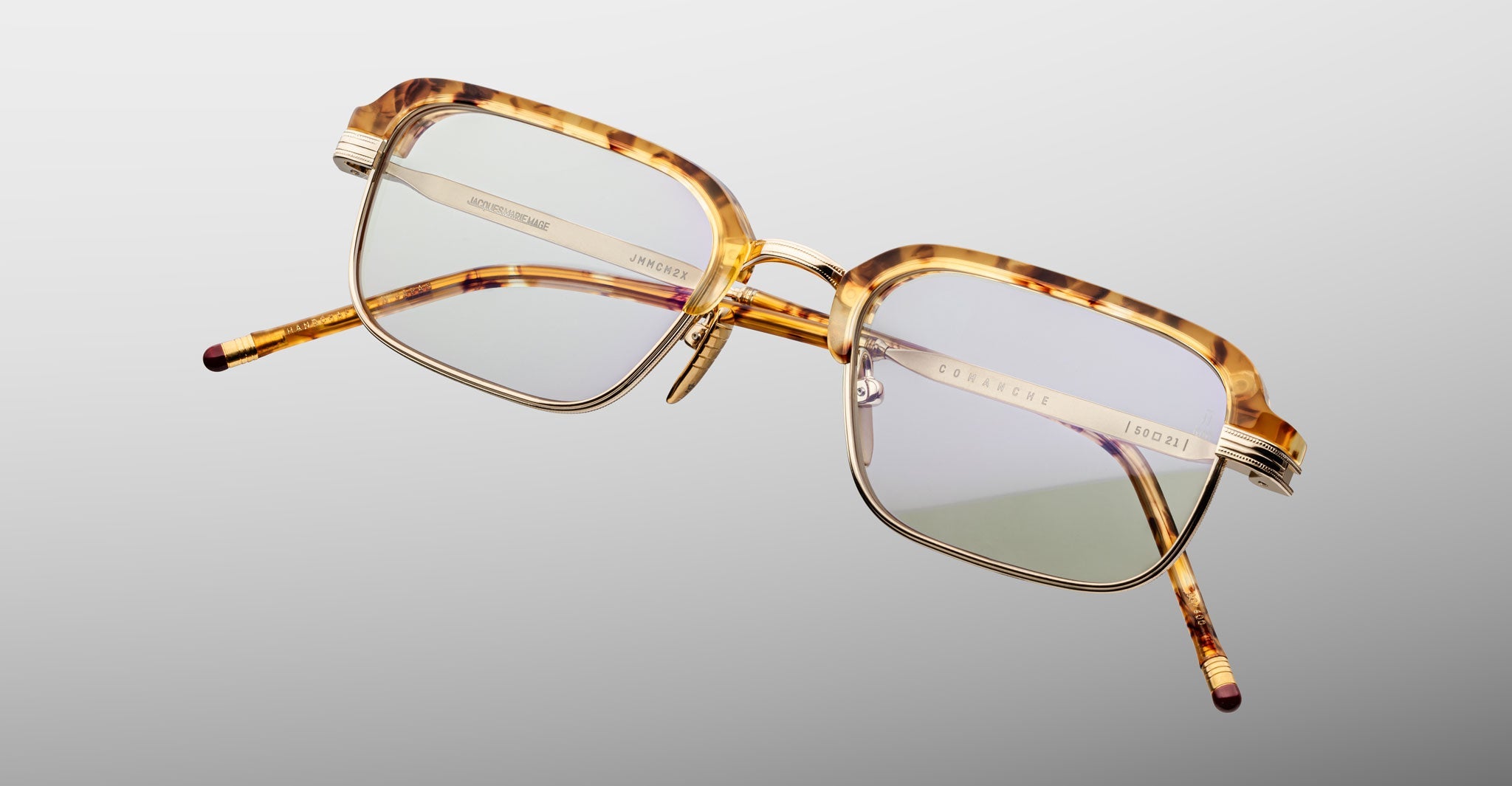 The Jacques Marie Mage Comanche eyeglasses feature rectangular tortoiseshell frames, thin gold metal accents, and clear lenses. This limited-edition eyewear by Jacques Marie Mage is shown on a plain light grey background.