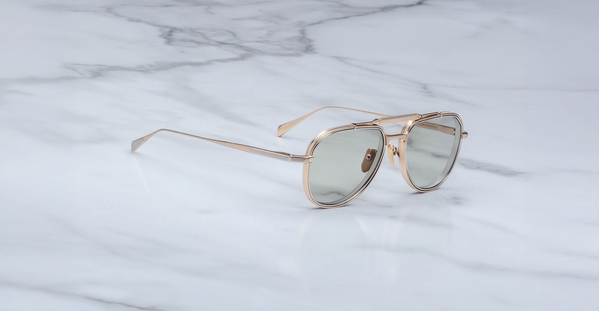 The Jacques Marie Mage Commander sunglasses, featuring gold frames and light gray lenses, rest on a white marble surface with gray veining.