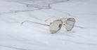 The Jacques Marie Mage Commander sunglasses, featuring gold frames and light gray lenses, rest on a white marble surface with gray veining.