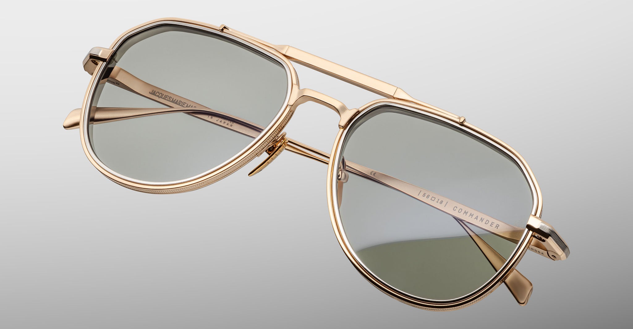 The Jacques Marie Mage Commander sunglasses feature gold metal aviator frames, gray-tinted lenses, a double bridge, and engraved branding on the inner arms, shown against a light gray background.
