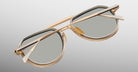 A close-up of Jacques Marie Mage Commander gold-framed sunglasses with gray lenses, folded on a light gradient background, showcasing the detailed frames and arm positioning.