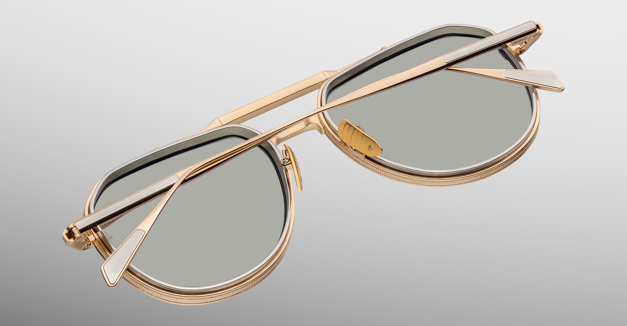 A close-up of Jacques Marie Mage Commander gold-framed sunglasses with gray lenses, folded on a light gradient background, showcasing the detailed frames and arm positioning.