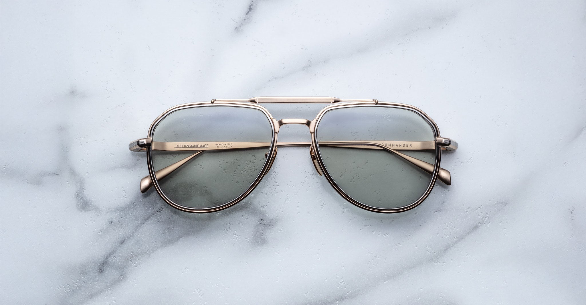 The Jacques Marie Mage Commander sunglasses, featuring gold frames and grey tinted lenses, rest on a white marble surface.