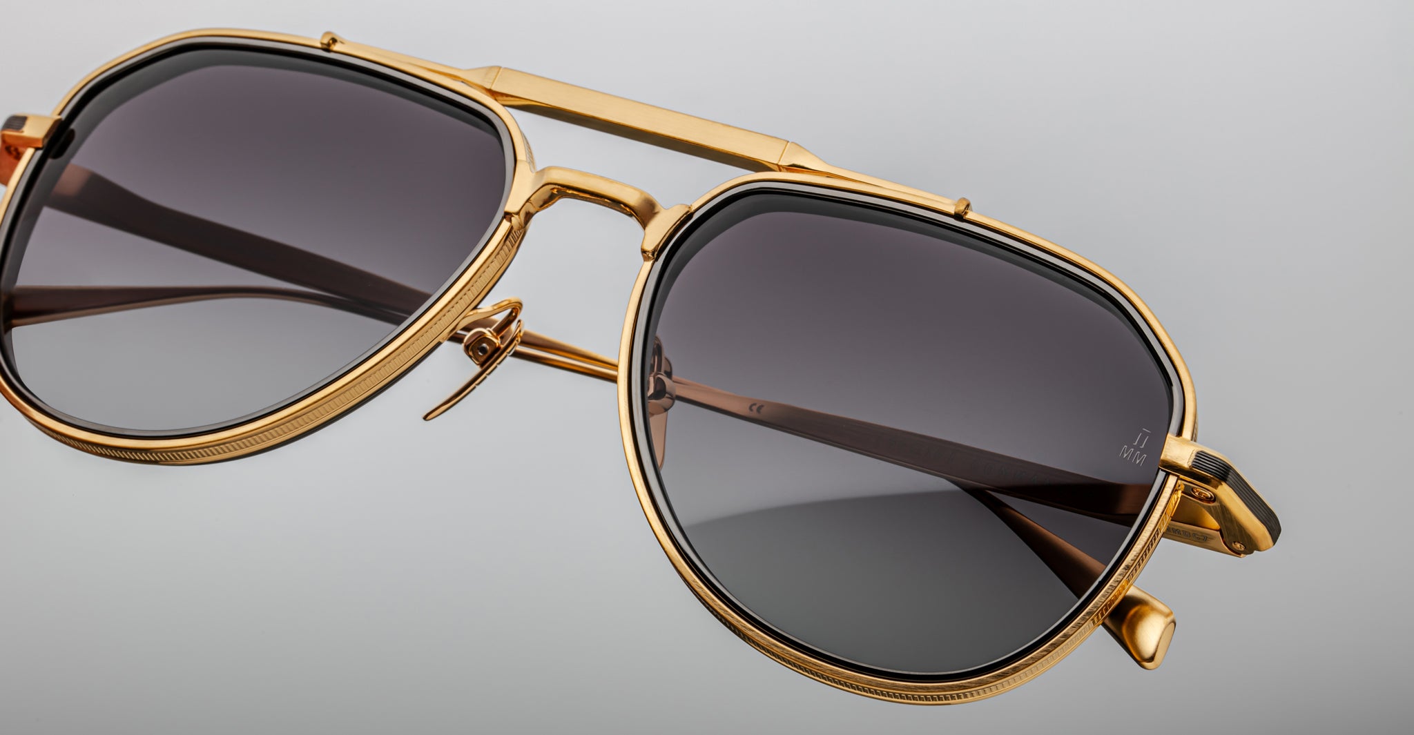 Close-up of Jacques Marie Mage Commander aviator sunglasses with gold frames, dark tinted lenses, a prominent top bar, and detailed metalwork on the frame edges, set against a plain light gray background.