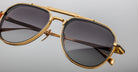 Close-up of Jacques Marie Mage Commander aviator sunglasses with gold frames, dark tinted lenses, a prominent top bar, and detailed metalwork on the frame edges, set against a plain light gray background.