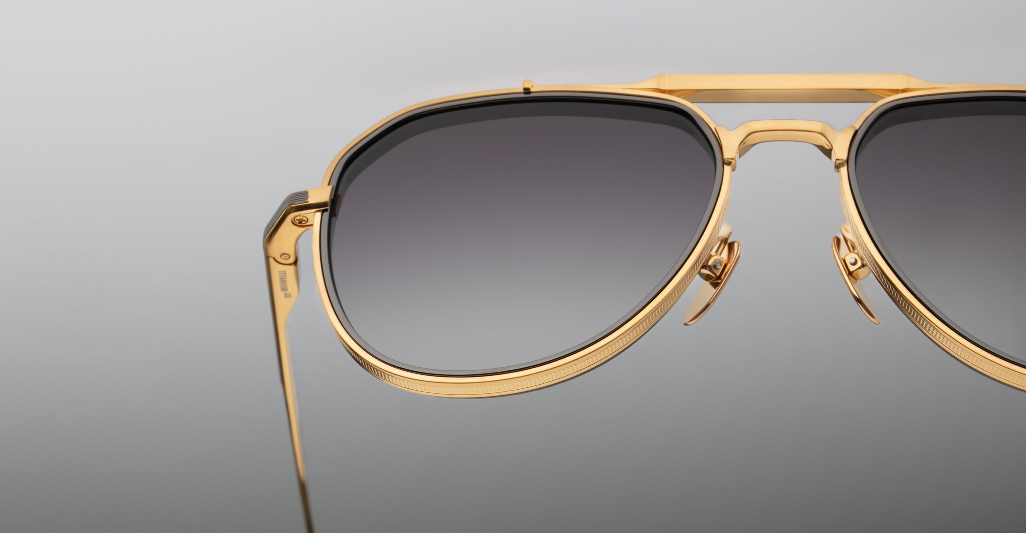 Close-up of Jacques Marie Mage Commander gold-framed aviator sunglasses with dark gradient lenses, set against a plain grey background. The image highlights the metallic details on the upper half of the frames.