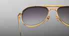 Close-up of Jacques Marie Mage Commander gold-framed aviator sunglasses with dark gradient lenses, set against a plain grey background. The image highlights the metallic details on the upper half of the frames.