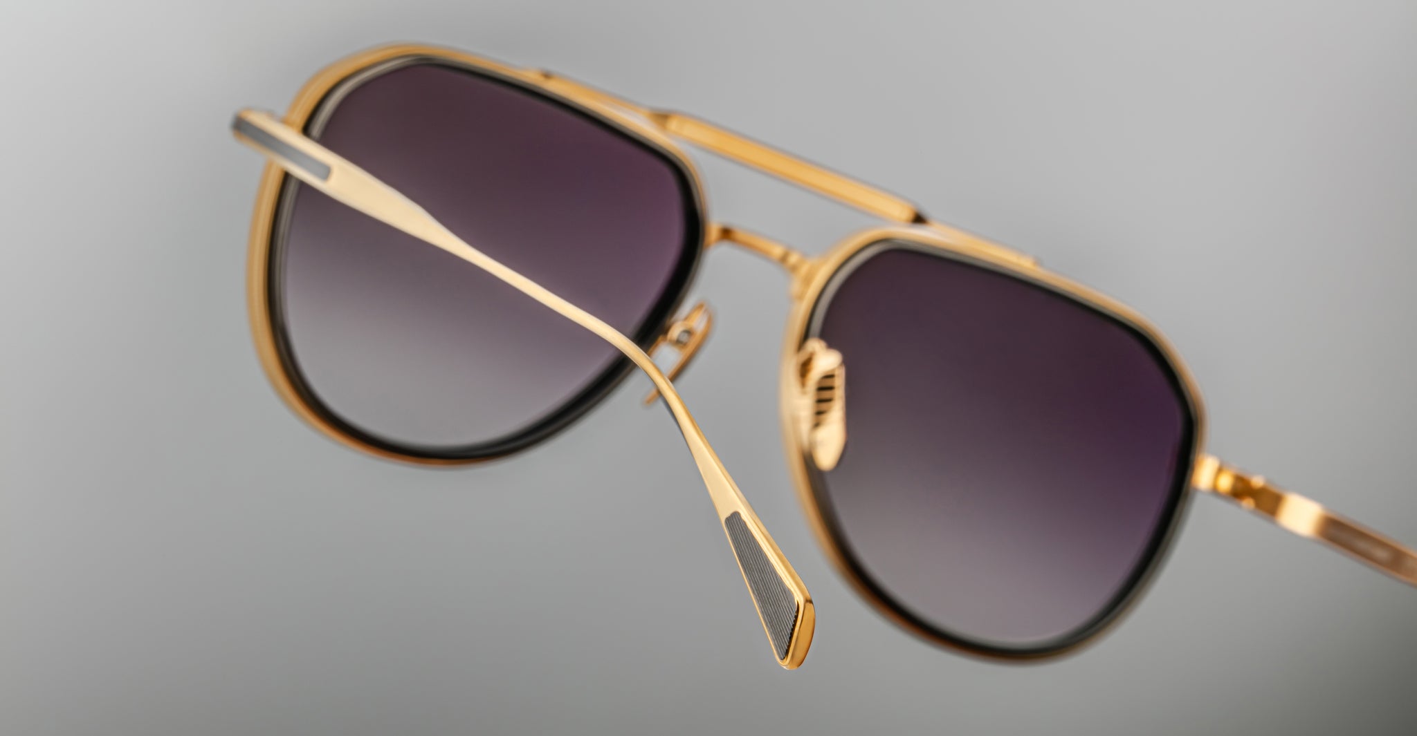 A close-up of Jacques Marie Mage Commander gold-framed aviator sunglasses with dark gradient lenses set against a plain gray background.