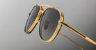 Close-up of Jacques Marie Mage Commander sunglasses with gold frames and dark lenses on a grey background, showcasing their detailed construction and reflective finish.