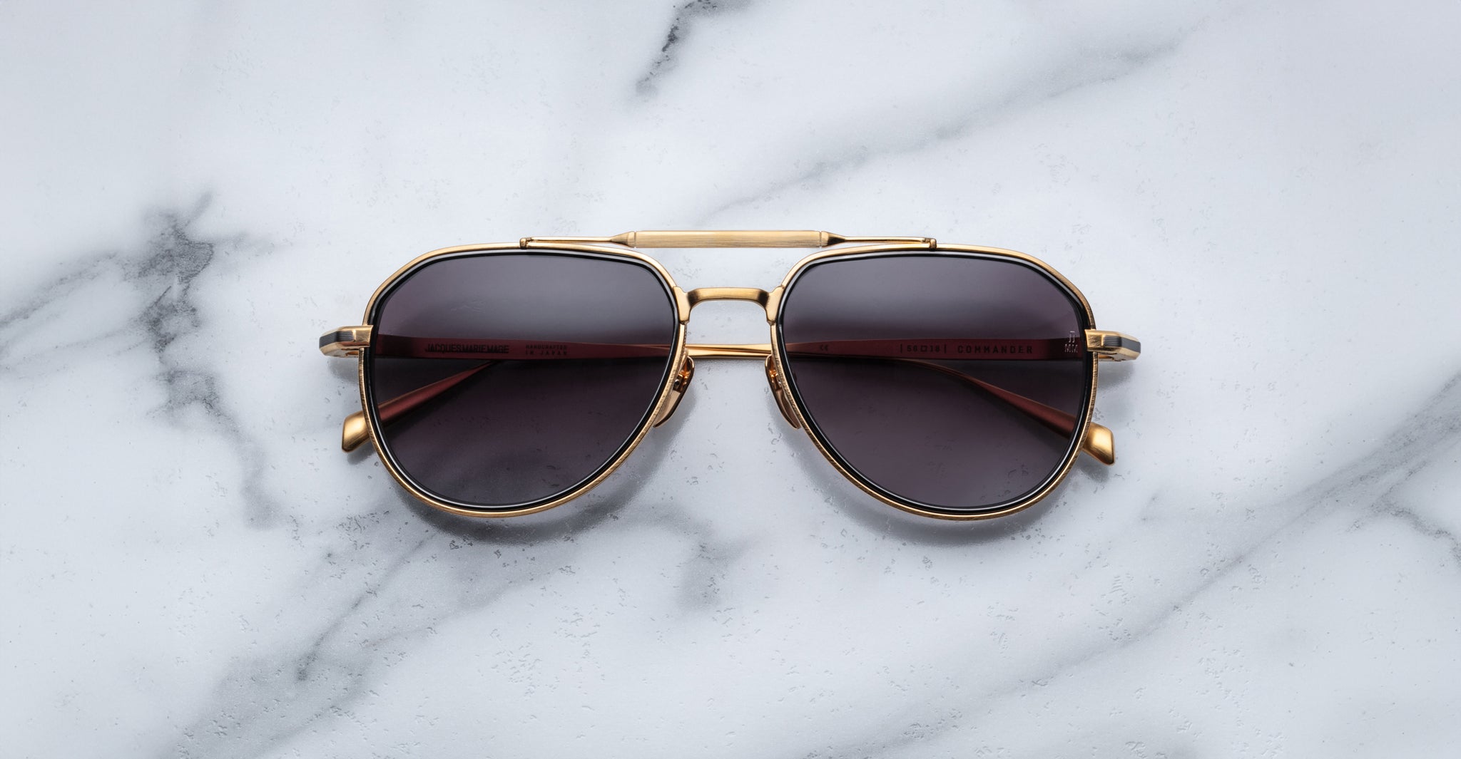 A pair of Jacques Marie Mage Commander gold-framed aviator sunglasses with dark lenses rests on a white marble surface with grey veining.