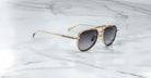 The Jacques Marie Mage Commander gold-framed aviator sunglasses with dark lenses rest at an angle on a white marble surface, set against a minimal background.