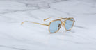 The Jacques Marie Mage Commander gold-framed aviator sunglasses with light blue lenses rest on a white marble surface adorned with gray veining.