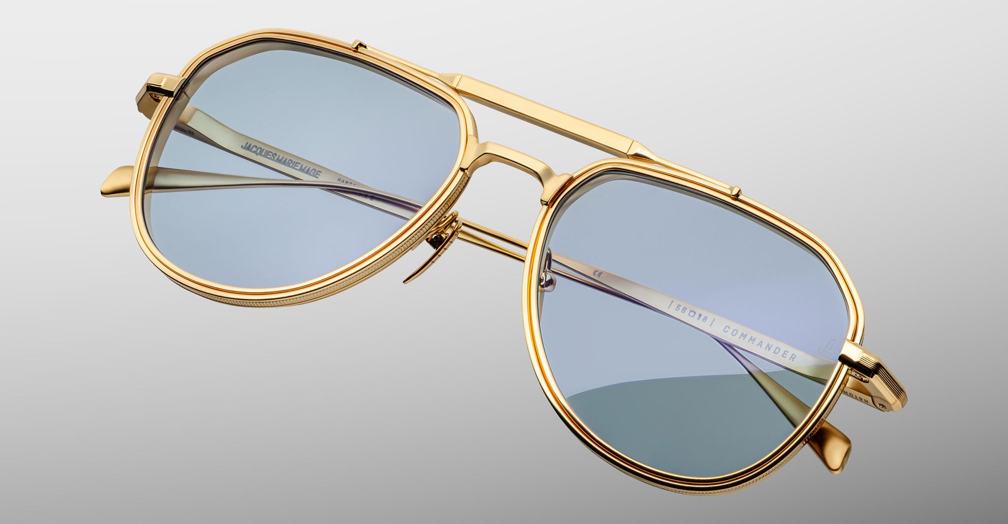 A pair of gold-framed aviator sunglasses with light blue tinted lenses, named Jacques Marie Mage Commander, is shown against a light gray background. The brand name is visible on the inside of the arm.